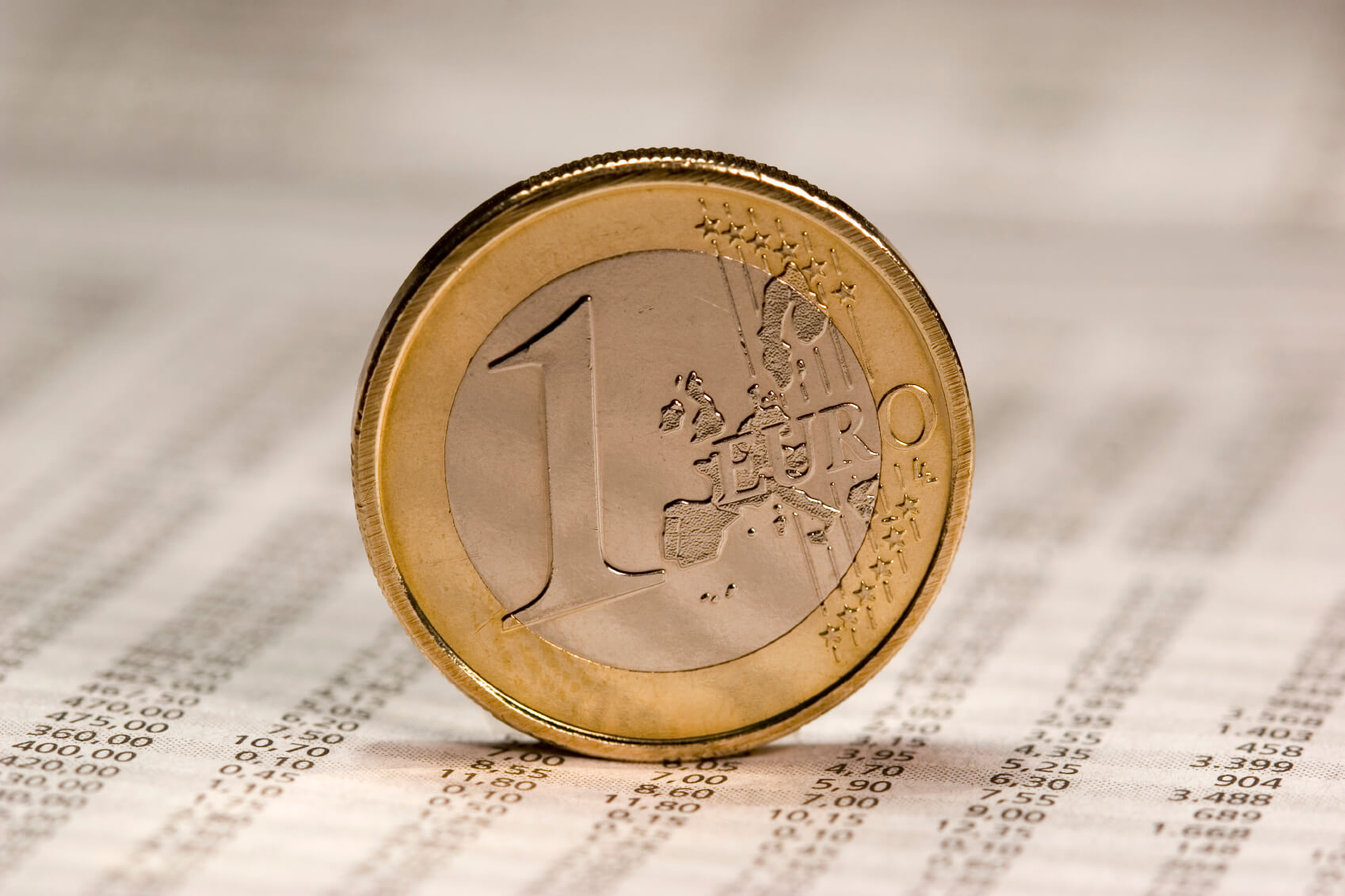 Eine 1-Euro-Münze steht hochkant auf einem Blatt Papier, auf dem Zahlen abgebildet sind