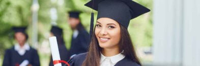Junge Absolventin steht vor einer Universität, im Hintergrund sind Graduierte zu sehen.